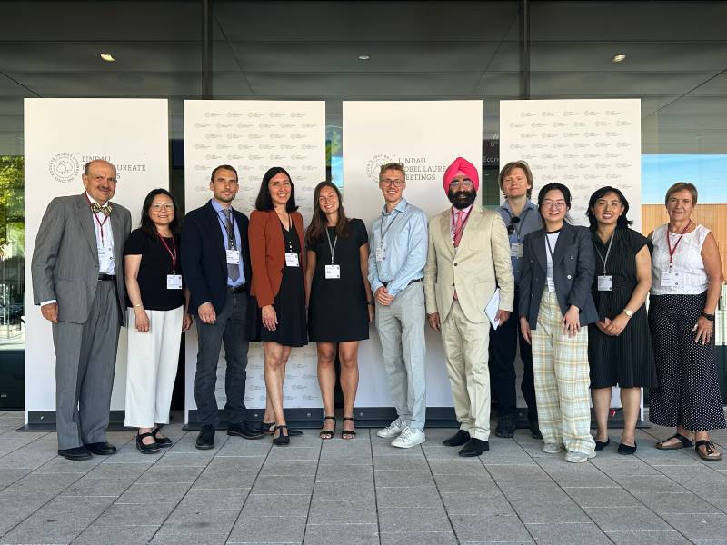 A diverse group of smiling people pose together in front of a board promoting the Lindau Foundation. 
